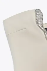 Vestido corto color crema, de escote asimétrico y sin mangas. Presenta una pieza metálica con apliques de strass en el escote.