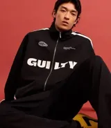 Campera deportiva negra con cuello alto y cierre frontal. Presenta franjas blancas contrastantes a lo largo de las mangas y detalles gráficos en blanco en el pecho, incluyendo la palabra "GUILTY" en grande.