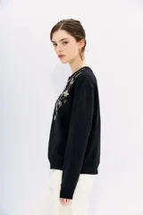 Buzo negro de algodón con cuello redondo y mangas largas. Presenta apliques bordados de piedras brillantes en forma de flores y otros motivos decorativos en el frente.