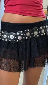 Cinturón ancho de cuero negro con apliques circulares metálicos plateados, cada uno con un diseño de sol grabado en el centro y rodeado de ojales metálicos.