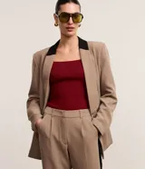 Blazer de tejido plano color beige con estructura de sastrería, cuello smoking con contraste negro en la nuca, botón único para cierre y solapas delanteras que simulan bolsillos.