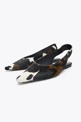 Zapato tipo bailarina destalonada en piel con pelo, con estampado animal print de vaca en blanco, marrón y negro. Tiene tira trasera con pieza elástica y acabado en punta. Suela plana de 1 cm de altura.