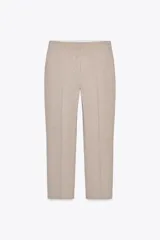 Pantalón de vestir color beige, de tiro bajo, corte recto y largo cropped. Cuenta con bolsillos laterales y traseros, cierre frontal con cremallera y gancho metálico, y abertura lateral en el bajo.