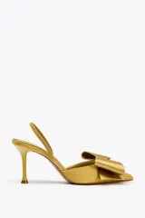Zapato de tacón destalonado color dorado, con efecto satinado, punta fina, tira trasera elástica y maxi lazo decorativo en la parte delantera.