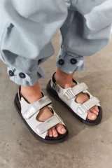 Sandalias de cuero color blanco con doble tira plateada con velcro, base negra y hebilla plateada.