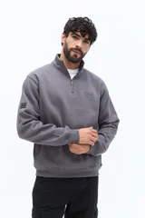 Buzo de algodón gris oscuro con cuello alto y cierre frontal parcial. Presenta un parche de marca en la manga izquierda y un logo bordado en el pecho.