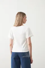 Remera de tejido acanalado color blanco, con cuello redondo y mangas cortas que presentan un detalle de terminación en puntilla en los bordes.