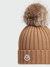 Gorro de lana de punto acanalado en color tostado, con un pompón de pelo en la parte superior y el parche con el logo de Moncler bordado en el frente del doblez.