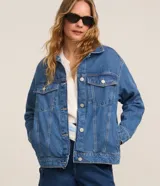 Campera trucker de jeans azul oscuro, corte amplio, con cuello clásico, cierre frontal con botones blancos, mangas largas y bolsillos delanteros con solapa.