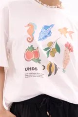 Remera blanca de algodón, corte oversize, con cuello redondo y manga corta. Presenta un estampado frontal con varias reposeras y hamacas de playa, y el texto "LA SIESTA".