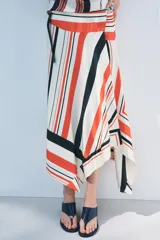 Falda midi de tiro medio con estampado de rayas rojas, negras y blancas. Tiene bajo asimétrico y cierre frontal tipo pareo ajustable con lazos negros.