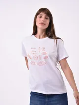 Remera blanca de algodón con cuello redondo y manga corta. Presenta un pequeño bordado en el pecho izquierdo con una figura que parece ser una persona acostada en una tabla de surf o flotador.