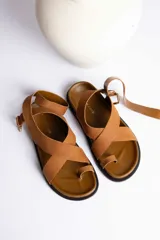 Sandalia plana de cuero color marrón topo, con tiras cruzadas en el empeine y tira sujetadora con hebilla dorada alrededor del tobillo. Posee una suela gruesa de goma negra.