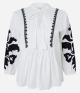 Blusa amplia de viscolino color blanco con bordados florales negros en mangas y escote, lazo en el cuello y mangas 3/4 con puño ajustado.