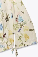 Camisa color crema con estampado de flores en tonos azules, amarillos y borgoña. Tiene cuello redondo con escote en pico, mangas largas con lazos y bajo ajustable con cordones.