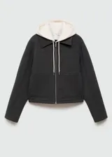 Chaqueta negra corta con capucha gris desmontable y ajustable con cordones. Presenta cuello camisero, cierre de cremallera doble, bolsillos laterales y bajo con goma elástica.