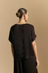 Top negro de manga corta con silueta amplia y caída fluida, confeccionado en un tejido liviano con leve brillo. Presenta un botón de nácar en la espalda con una pequeña abertura y mangas amplias tipo kimono.