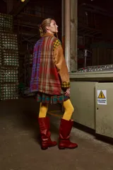 Chaqueta con cuerpo de lana a cuadros en tonos marrón, verde y amarillo. Mangas y cuello de cuero agamuzado color camel. Cierre frontal con cremallera. Falda corta a cuadros escoceses en tonos verde, rojo y azul. Medias amarillas y botas de cuero marrón. 