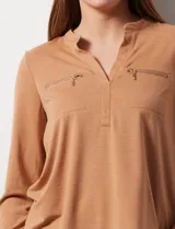 Blusa blanca de poliéster y elastano, con cuello a la base, escote en V y mangas tres cuartos. Presenta dos bolsillos frontales con cierre metálico dorado.