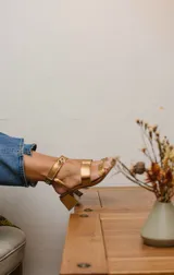 Sandalias plateadas con dos tiras horizontales sobre el pie y una tira vertical que se une a una pulsera con hebilla en el tobillo. La pulsera tiene ojales y tachas combinadas. El taco es cuadrado y de 5 cm.