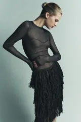 Vestido midi negro, semitransparente, de manga larga y cuello redondo. La parte superior presenta un tejido de punto fino con costuras que simulan un bustier. La falda está compuesta por capas de flecos largos y densos.