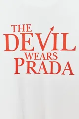 Camiseta blanca de algodón con cuello redondo, mangas cortas y hombros caídos. Estampado frontal con el título de la película "El Diablo Viste de Prada" en letras rojas.