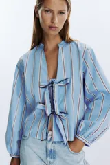 Camisa celeste con rayas verticales marrones y blancas, cuello redondo con escote en pico y mangas largas abullonadas. Cierre frontal con lazos con pespuntes marcados.