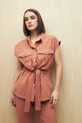 Chaleco color beige con cuello camisero, sin mangas, cierre frontal con botones y cinturón de lazo. Presenta bolsillos de parche en el pecho y pinzas verticales en el frente.