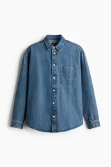Camisa de denim azul de algodón rígido, corte holgado, cuello inglés, tapeta clásica con botones blancos, bolsillo de parche en el pecho, canesú en la espalda, mangas largas con puños abotonados y bajo levemente redondeado.