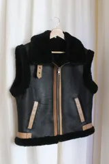 Chaleco negro de cuero con cuello y borde inferior de piel sintética negra. Cierre frontal con cremallera color cobre y detalles en cuero color tostado.