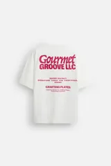 Camiseta blanca de corte relajado, con cuello redondo y mangas cortas. Presenta estampados combinados en contraste en la parte delantera y trasera con la frase "Gourmet Groove LLC".