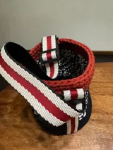 Bolso tejido a crochet con hilo de trapillo en colores negro, blanco y rojo. Tiene base de cuero vegano y correa de tela ajustable con detalles en rojo y blanco.