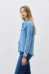 Camisa de denim celeste con cuello clásico, cierre frontal con botones a presión, dos bolsillos con solapa en el pecho y mangas largas con puños abotonados.