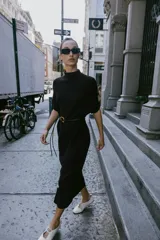 Vestido negro de tejido de punto, corte midi y silueta holgada, con cuello alto acanalado y mangas cortas tipo murciélago. Se ajusta a la cintura con un cinturón fino de efecto piel negro con hebilla dorada en forma de C.