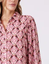 Camisa de gasa con cuello con solapa y apertura central con botones. Presenta un estampado de patrones geométricos en tonos rosados, marrones y crema, con un borde inferior en un patrón geométrico más denso en tonos rojizos/rosados. Las mangas son amplias de largo tres cuartos con puño elastizado.