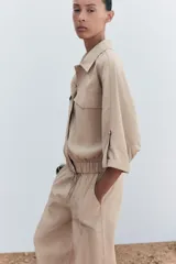 Camisa color beige de cuello solapa y manga corta con trabilla y botón. Tiene bolsillos delanteros de plastrón con solapa, bajo con elástico y cierre frontal con botones a presión.