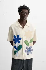 Camisa de manga corta color crudo, con cuello bowling, corte relaxed fit y cierre frontal con botones. Presenta apliques de parches con texturas combinadas a contraste en el delantero y aberturas laterales en el bajo.