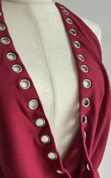 Vestido corto ajustado color rojo oscuro, estilo halter con escote pronunciado en V que se ajusta con ojales metálicos a lo largo del borde. El ruedo también presenta ojales metálicos.