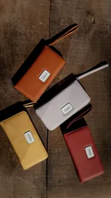 Billetera larga tipo clutch, color negro, con cierre perimetral y correa de mano desmontable. Presenta placa metálica con logo en el frente.