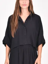 Camisa negra de manga corta con cuello camisero y botones frontales. Presenta un corte cropped y textura acanalada o plisada.