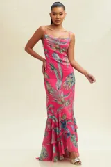Vestido largo de tirantes finos, con escote drapeado y estampado de hojas tropicales en tonos verdes y azules sobre fondo fucsia. Tiene corte al bies y volantes en el ruedo.