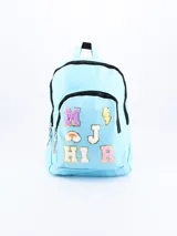 Mochila celeste con ribetes negros, asa superior y correas acolchadas ajustables. Decorada con parches de letras y símbolos en colores brillantes y texturas variadas.