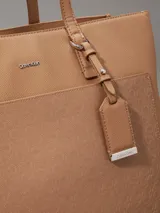 Cartera tote Calvin Klein color tostado, de tamaño mediano, confeccionada en cuero sintético granulado con textura de logo monograma en el bolsillo delantero. Tiene asas fijas, cierre magnético, bolsillo exterior, compartimento interior con cremallera y colgante extraíble con logo.