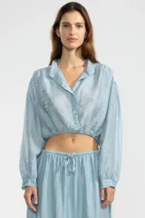 Blusa corta celeste en tejido arrugado liviano, con cuello mao, botones al frente y mangas abullonadas. Terminada con ruedo elastizado.