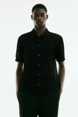 Camisa negra de manga corta con tejido calado tipo crochet, cuello inglés, botones frontales y corte recto.