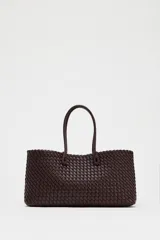 Bolso shopper alargado color marrón chocolate con diseño trenzado. Tiene doble asa de hombro y cierre superior con botón imantado. Incluye una bolsa interior con cierre fruncido.