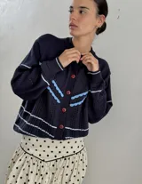 Saco de punto calado color beige claro, con cuello redondo y cierre frontal de botones azules. Presenta detalles decorativos en crochet verde y negro en el frente y puños.