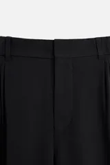 Pantalón negro de corte amplio confeccionado en tejido con mezcla de viscosa. Presenta cintura elástica, pliegues en la parte delantera, bolsillos laterales y traseros de vivo. Cierre frontal con cremallera y gancho metálico oculto.