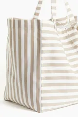 Bolso de playa tipo tote bag, de lona de algodón a rayas horizontales color beige y blanco. Tiene doble asa de hombro y un compartimento interior.