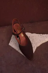 Sandalias Agnes de gamuza negra con punta fina y detalle metálico dorado en la puntera. Tira delantera y tira al tobillo con hebilla dorada.